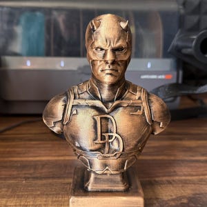 May include: A bronze-colored bust of a superhero with a horned mask and the letter "D" on the chest. The bust is on a rectangular base and appears to be a collectible item or decorative piece.