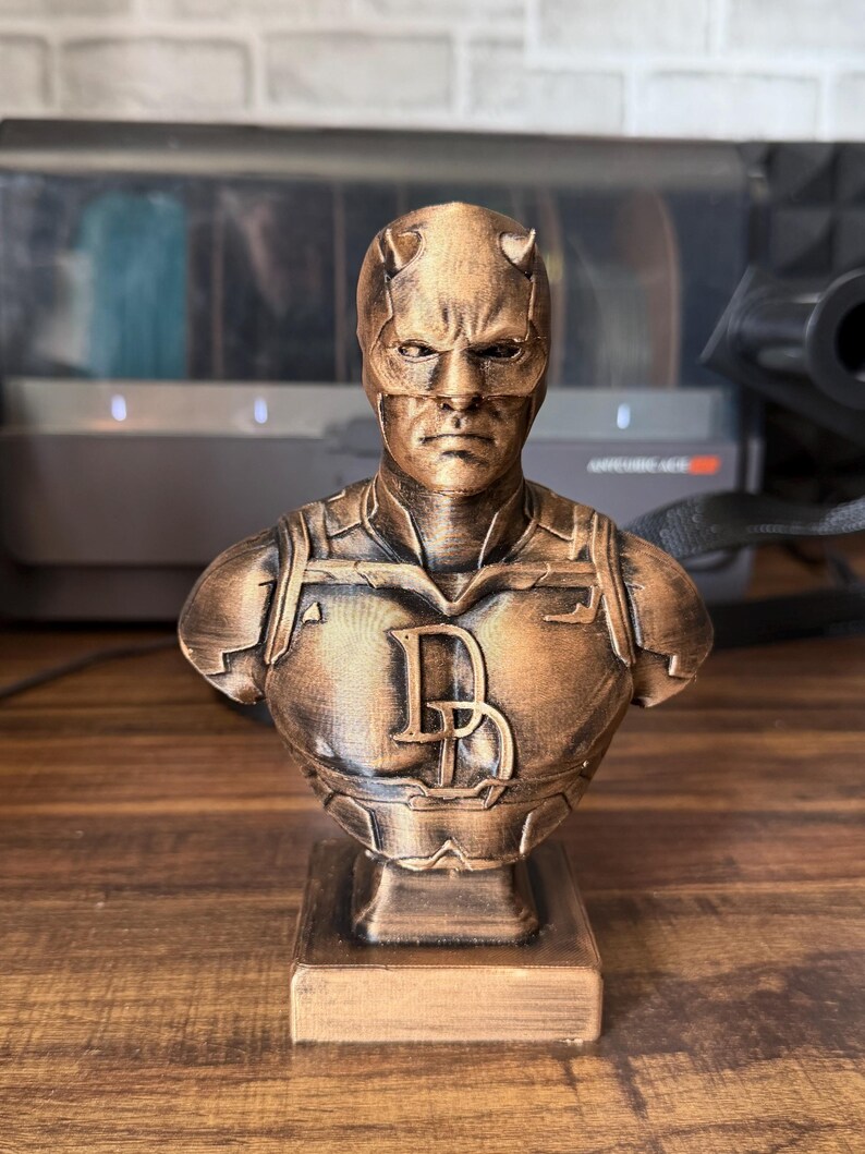 May include: A bronze-colored bust of a superhero with a horned mask and the letter "D" on the chest. The bust is on a rectangular base and appears to be a collectible item or decorative piece.
