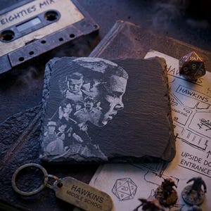 May include: A dark gray slate coaster featuring an etched portrait of a person and other figures. A vintage cassette tape labeled "EIGHTIES MIX" and a "Hawkins Middle School" keychain are also visible, along with a map and dice.