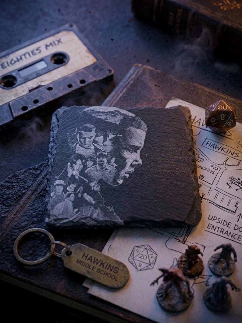 May include: A dark gray slate coaster featuring an etched portrait of a person and other figures. A vintage cassette tape labeled "EIGHTIES MIX" and a "Hawkins Middle School" keychain are also visible, along with a map and dice.