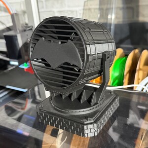 以下が含まれることがあります： 黒い3Dプリントのバットシグナルスポットライトのレプリカ。 スポットライトは、中央にコウモリのシンボルが付いた円形のデザインで、正方形のベースに取り付けられています。 デザインには、前面に格子状のパターンが含まれています。