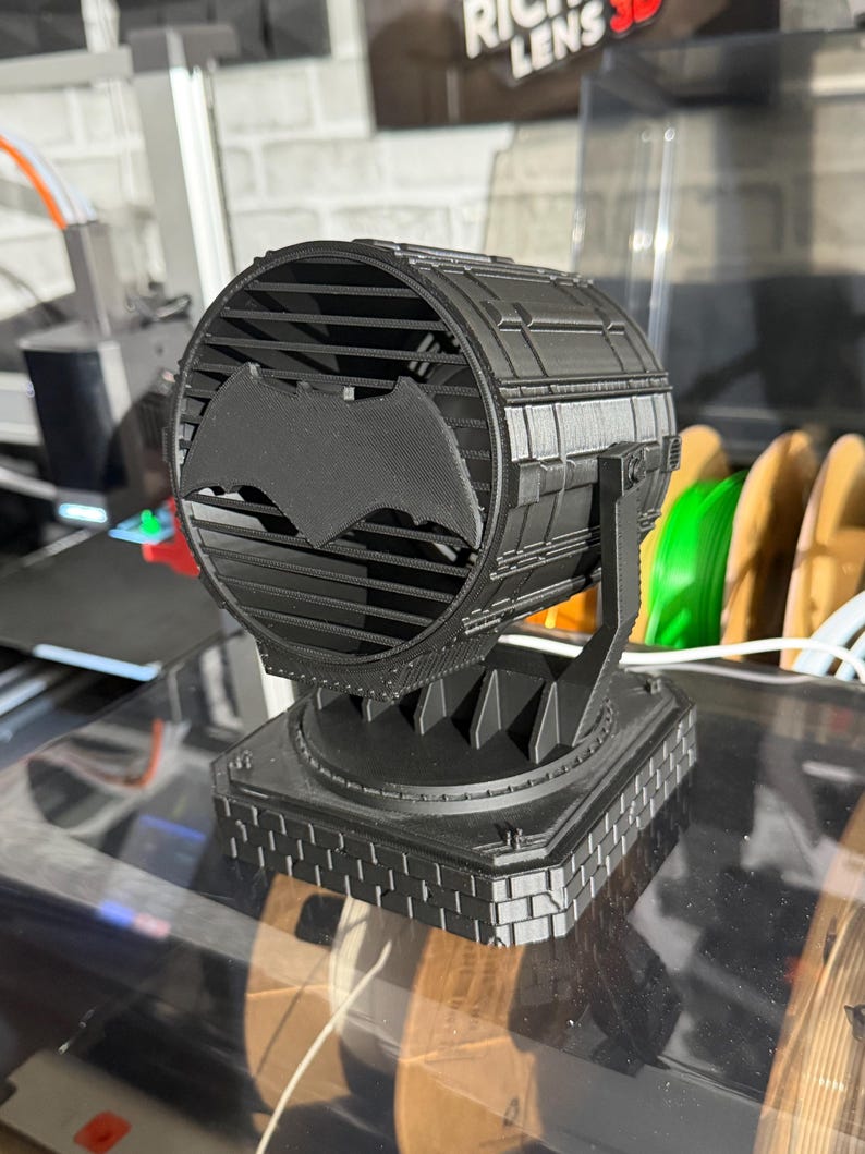 以下が含まれることがあります： 黒い3Dプリントのバットシグナルスポットライトのレプリカ。 スポットライトは、中央にコウモリのシンボルが付いた円形のデザインで、正方形のベースに取り付けられています。 デザインには、前面に格子状のパターンが含まれています。