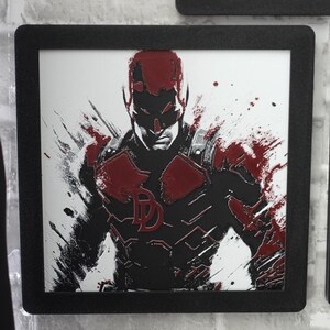 May include: A square framed wall art piece featuring a stylized illustration of the character Daredevil. The artwork uses a high-contrast black and white palette with bold red accents, showcasing a paint-splatter effect and the iconic DD logo.