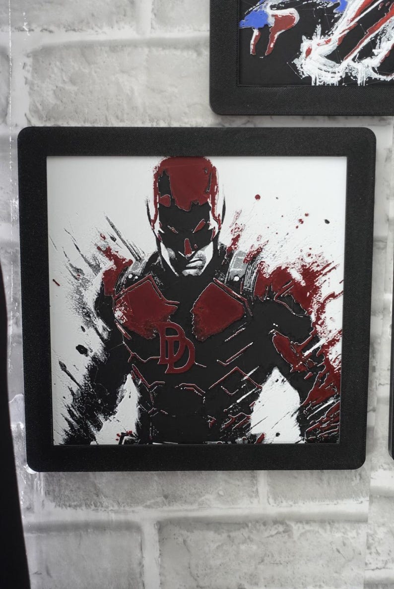 May include: A square framed wall art piece featuring a stylized illustration of the character Daredevil. The artwork uses a high-contrast black and white palette with bold red accents, showcasing a paint-splatter effect and the iconic DD logo.