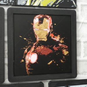 May include: A square framed wall art piece featuring a stylized graphic illustration of Iron Man in red and gold tones against a solid black background. The artwork displays a distressed, paint-splatter effect with a glowing white chest arc reactor.