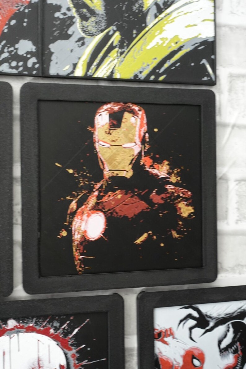 May include: A square framed wall art piece featuring a stylized graphic illustration of Iron Man in red and gold tones against a solid black background. The artwork displays a distressed, paint-splatter effect with a glowing white chest arc reactor.