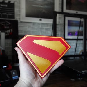 May include: A diamond-shaped emblem featuring the iconic Superman logo design. The object is constructed with a vibrant red outer border and a central red field, accented by a contrasting metallic gold finish on the raised letter S and inner border.