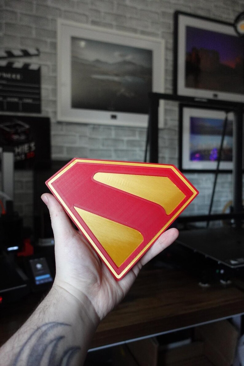 May include: A diamond-shaped emblem featuring the iconic Superman logo design. The object is constructed with a vibrant red outer border and a central red field, accented by a contrasting metallic gold finish on the raised letter S and inner border.