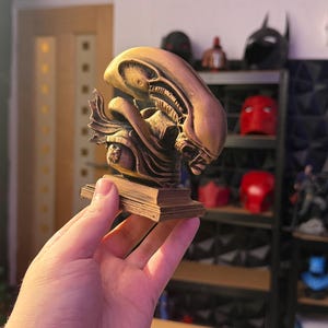 May include: A bronze-colored Alien bust sculpture, featuring intricate details of the creature's head and face. The sculpture is mounted on a rectangular base. Shelves in the background display other superhero-themed items.