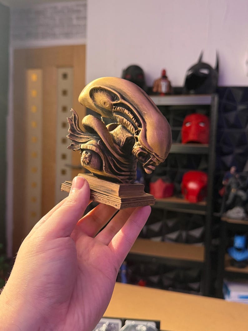 May include: A bronze-colored Alien bust sculpture, featuring intricate details of the creature's head and face. The sculpture is mounted on a rectangular base. Shelves in the background display other superhero-themed items.