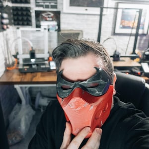 May include: A red and black mask with a textured finish. The mask has a distinctive design with a bat-like emblem on the forehead and eye openings. The mask is held up by a person.