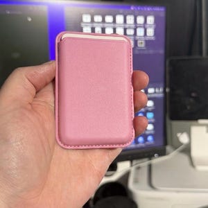May include: A pink leather card holder, held in a hand. The rectangular case has a smooth surface and a contrasting white interior. The edges are neatly stitched, and the overall design is minimalist and sleek.
