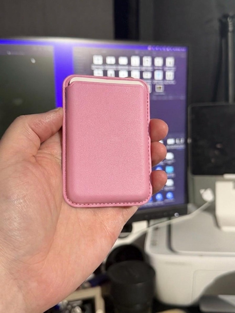 May include: A pink leather card holder, held in a hand. The rectangular case has a smooth surface and a contrasting white interior. The edges are neatly stitched, and the overall design is minimalist and sleek.