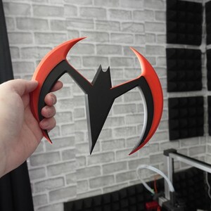 May include: A crescent-shaped throwing weapon featuring a central black bat silhouette integrated into a curved frame. The outer edges of the frame are bright orange, while the inner section is black, creating a sharp, aerodynamic design.