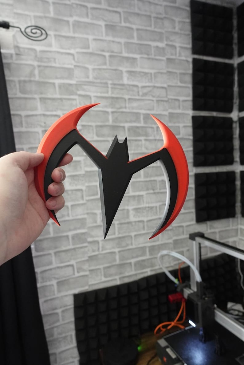 May include: A crescent-shaped throwing weapon featuring a central black bat silhouette integrated into a curved frame. The outer edges of the frame are bright orange, while the inner section is black, creating a sharp, aerodynamic design.