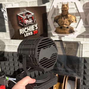 以下が含まれることがあります： 黒い3Dプリントのバットマンのスポットライト。象徴的なバットのシンボルが付いています。 スポットライトは、レンガのようなデザインの長方形のベースに取り付けられています。 画像には、バットマンの胸像と「Richie's Lens 3D」と書かれたサインも表示されています。
