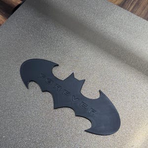 May include: A black, flat, bat-shaped emblem featuring the word FOREVER embossed in capital letters across the center. The object has a matte finish and a distinct wing-like silhouette with pointed edges, crafted with a visible layered texture.