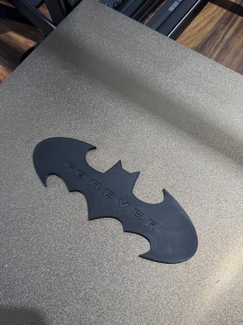 May include: A black, flat, bat-shaped emblem featuring the word FOREVER embossed in capital letters across the center. The object has a matte finish and a distinct wing-like silhouette with pointed edges, crafted with a visible layered texture.