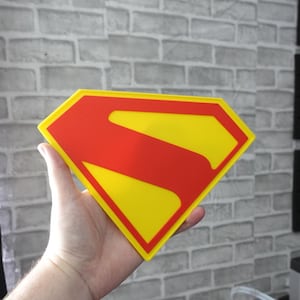 May include: A diamond-shaped sign featuring the iconic Superman logo with a bright yellow background and a bold red border and central S-shaped emblem. The flat, graphic design is rendered in vibrant primary colors, creating a clean and recognizable superhero symbol.