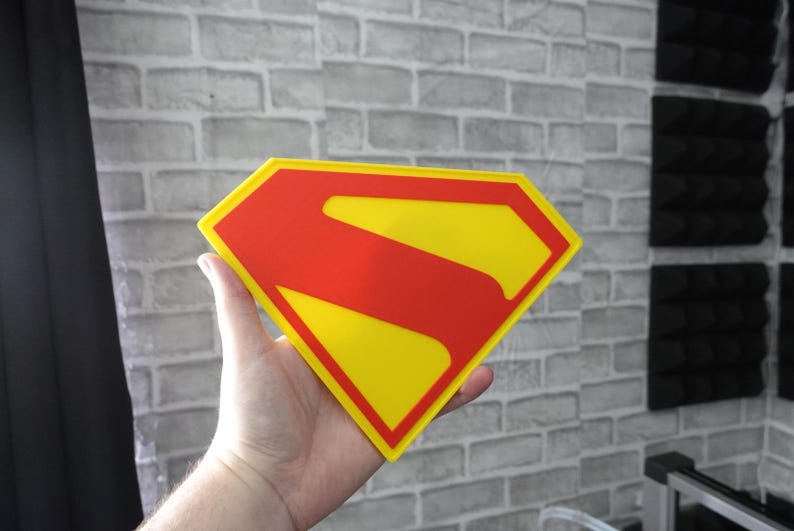 May include: A diamond-shaped sign featuring the iconic Superman logo with a bright yellow background and a bold red border and central S-shaped emblem. The flat, graphic design is rendered in vibrant primary colors, creating a clean and recognizable superhero symbol.