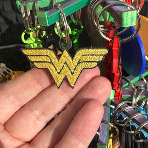 K&ouml;nnte beinhalten: Ein gold-schwarzer Wonder Woman Logo-Schl&uuml;sselanh&auml;nger wird vor einer Pr&auml;sentation von Schl&uuml;sselanh&auml;ngern gehalten. Das Logo ist mit goldenem Faden bestickt und hat einen schwarzen Rand. Andere Schl&uuml;sselanh&auml;nger im Hintergrund sind in verschiedenen Farben und Formen.