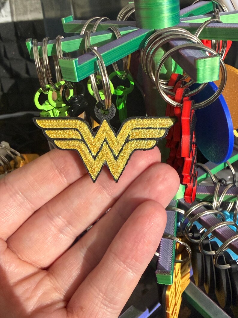K&ouml;nnte beinhalten: Ein gold-schwarzer Wonder Woman Logo-Schl&uuml;sselanh&auml;nger wird vor einer Pr&auml;sentation von Schl&uuml;sselanh&auml;ngern gehalten. Das Logo ist mit goldenem Faden bestickt und hat einen schwarzen Rand. Andere Schl&uuml;sselanh&auml;nger im Hintergrund sind in verschiedenen Farben und Formen.