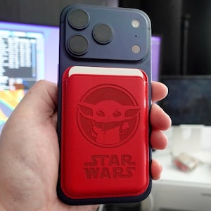 May include: A blue smartphone with a red cardholder featuring a Grogu design and the words "STAR WARS". The phone has three camera lenses. The cardholder is attached to the back of the phone.
