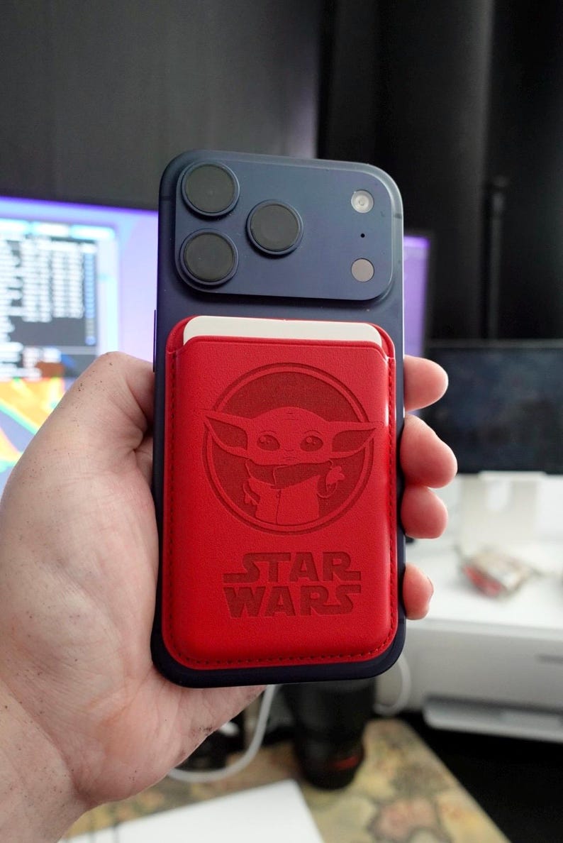 May include: A blue smartphone with a red cardholder featuring a Grogu design and the words "STAR WARS". The phone has three camera lenses. The cardholder is attached to the back of the phone.