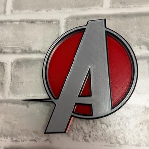 May include: A three-dimensional emblem featuring a large silver letter A centered within a red circular background, enclosed by a thin black circular border. The design includes a horizontal line extending from the left side of the letter A.