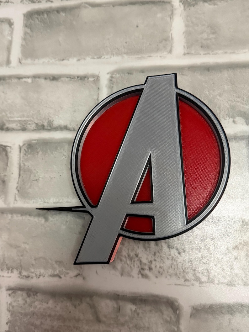 May include: A three-dimensional emblem featuring a large silver letter A centered within a red circular background, enclosed by a thin black circular border. The design includes a horizontal line extending from the left side of the letter A.
