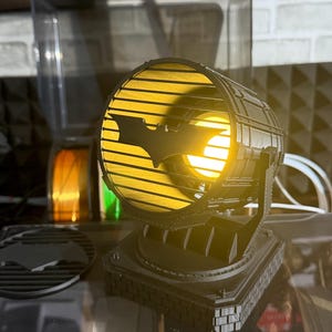 以下が含まれることがあります： 黒い3Dプリントのバットマンのスポットライトランプ。黄色い光を発し、バットのシンボルと工業的なデザインが特徴です。透明な表面に置かれ、サーチライトに似ています。