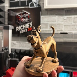 May include: A gold-colored 3D-printed figurine of a cartoon dog, posed on a circular base. The dog has a collar and a friendly expression. The figurine is held in a hand, with a blurred background including a sign that says "Richie's Lens 3D".