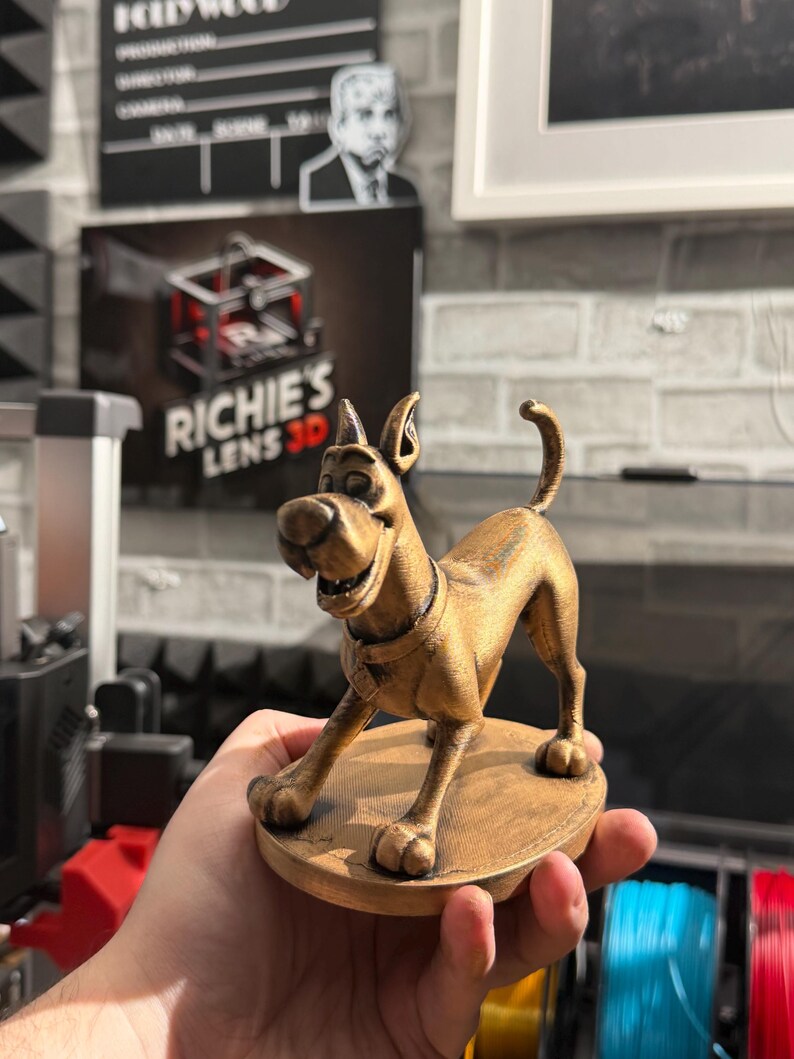 May include: A gold-colored 3D-printed figurine of a cartoon dog, posed on a circular base. The dog has a collar and a friendly expression. The figurine is held in a hand, with a blurred background including a sign that says "Richie's Lens 3D".
