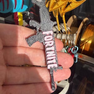 K&ouml;nnte beinhalten: Ein grau-rosa Fortnite-Schl&uuml;sselanh&auml;nger. Der Schl&uuml;sselanh&auml;nger hat die Form einer Waffe mit dem Wort "FORTNITE" in rosa Buchstaben. Im Hintergrund sind weitere Schl&uuml;sselanh&auml;nger mit blauen, goldenen und roten Designs zu sehen.