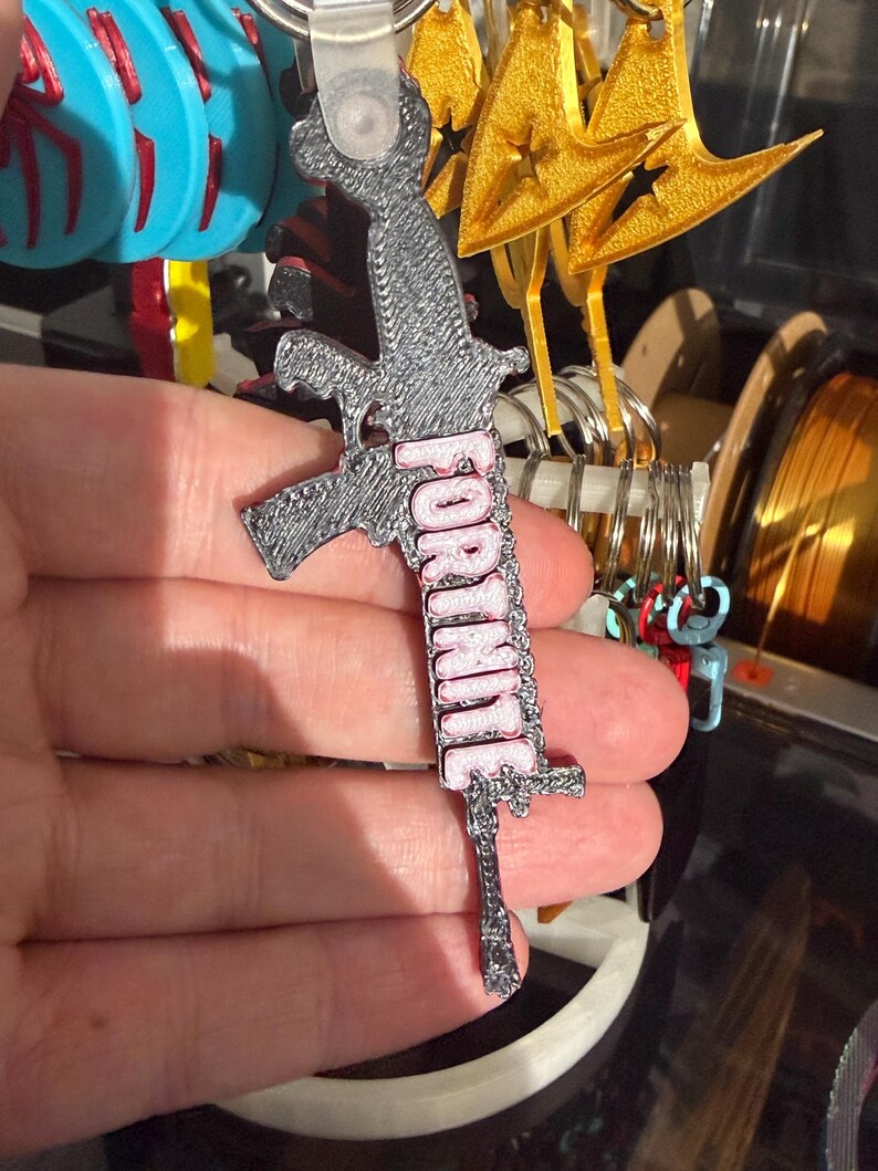 K&ouml;nnte beinhalten: Ein grau-rosa Fortnite-Schl&uuml;sselanh&auml;nger. Der Schl&uuml;sselanh&auml;nger hat die Form einer Waffe mit dem Wort "FORTNITE" in rosa Buchstaben. Im Hintergrund sind weitere Schl&uuml;sselanh&auml;nger mit blauen, goldenen und roten Designs zu sehen.