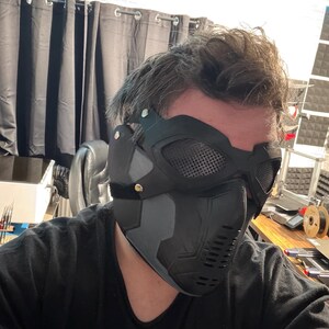 May include: A black and gray tactical mask with mesh eye coverings and a vented lower face section. The mask has a matte finish and is secured with a black strap. The design suggests a futuristic or military theme.