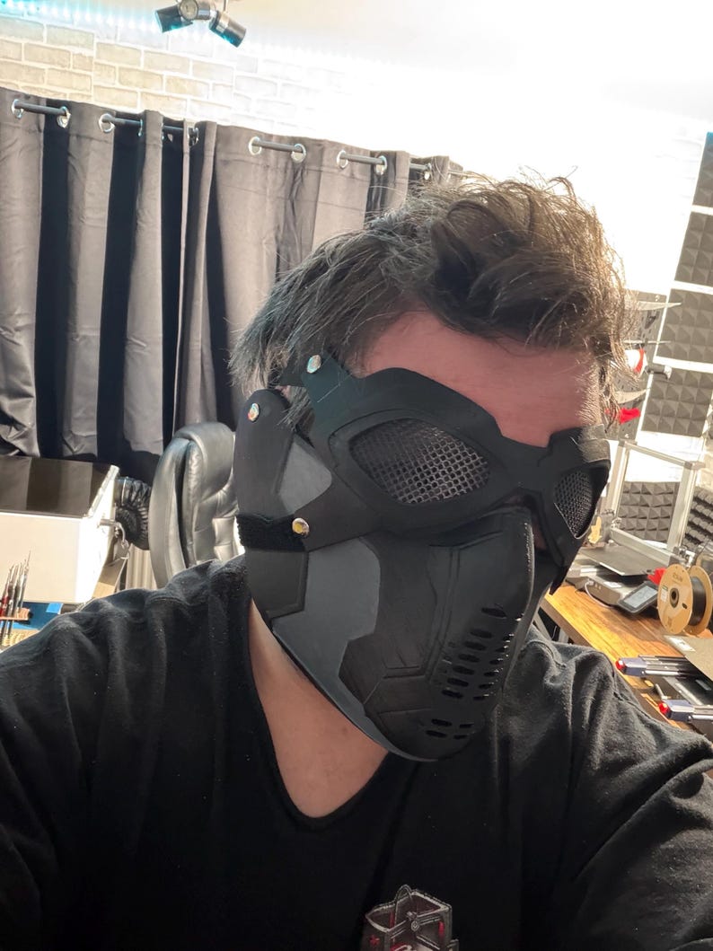 May include: A black and gray tactical mask with mesh eye coverings and a vented lower face section. The mask has a matte finish and is secured with a black strap. The design suggests a futuristic or military theme.