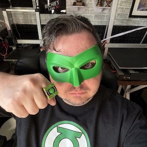 Puede incluir: Una persona con una m&aacute;scara verde y una camiseta negra con un gran logotipo verde y blanco de Green Lantern. La persona tambi&eacute;n lleva un anillo dorado con un emblema verde. El fondo incluye una pared de ladrillo y varios objetos decorativos.