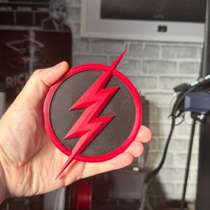 May include: A red and black circular emblem featuring a red lightning bolt. The emblem is held in a hand. The lightning bolt is the central design element, set against a black circular background, with a red border.
