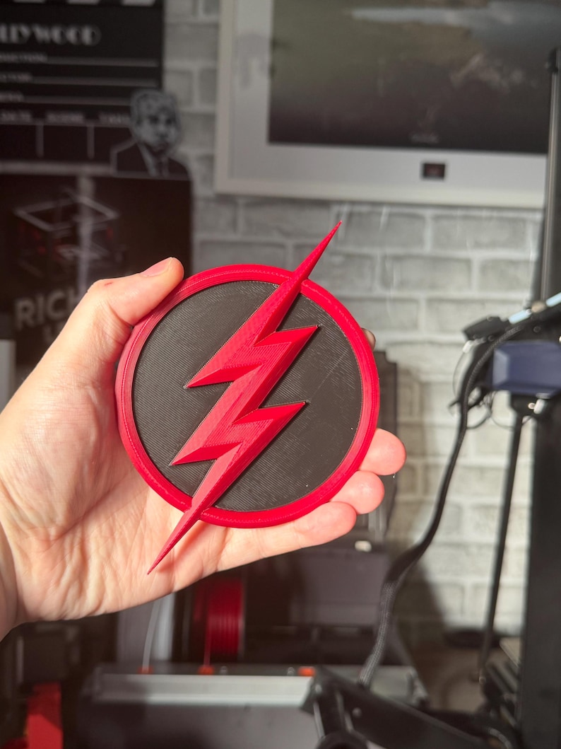 May include: A red and black circular emblem featuring a red lightning bolt. The emblem is held in a hand. The lightning bolt is the central design element, set against a black circular background, with a red border.