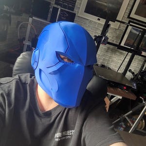 May include: A vibrant blue helmet with a futuristic design, featuring sharp angles and eye openings. The person in the image is wearing a black t-shirt with the text "PURE PROJECTS NEW YORK CITY". The helmet has ear-like protrusions.