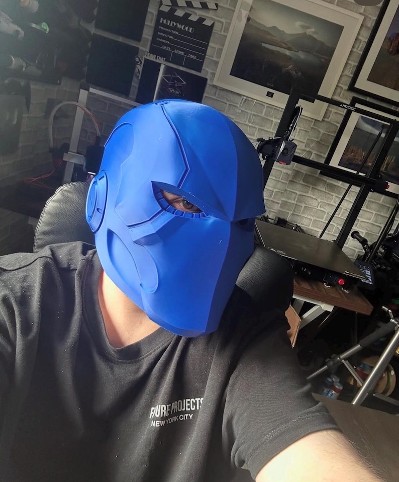May include: A vibrant blue helmet with a futuristic design, featuring sharp angles and eye openings. The person in the image is wearing a black t-shirt with the text "PURE PROJECTS NEW YORK CITY". The helmet has ear-like protrusions.