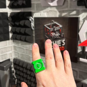 Puede incluir: Se muestra un anillo verde brillante con un dise&ntilde;o circular y un marco cuadrado. El anillo se lleva en un dedo. El fondo incluye una pared de ladrillos y un letrero que dice "HIE'S 3D".