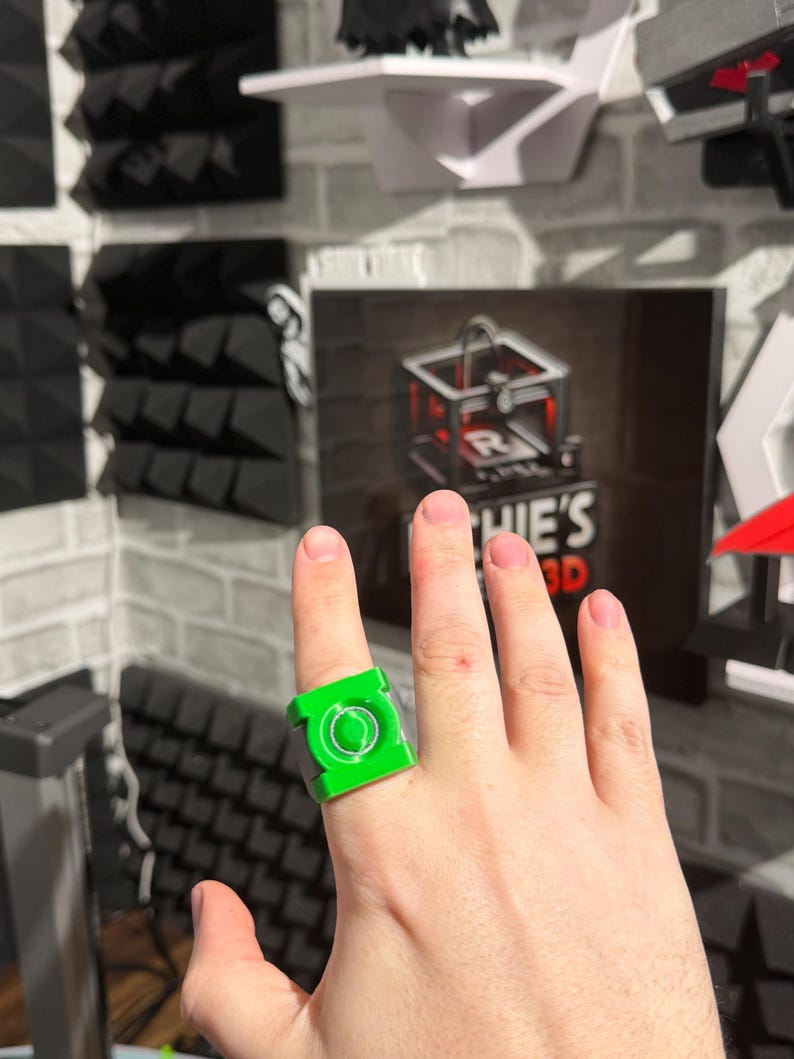 Puede incluir: Se muestra un anillo verde brillante con un dise&ntilde;o circular y un marco cuadrado. El anillo se lleva en un dedo. El fondo incluye una pared de ladrillos y un letrero que dice "HIE'S 3D".