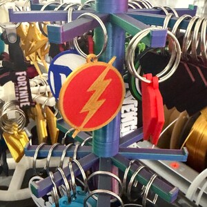 K&ouml;nnte beinhalten: Ein mehrfarbiger Ausstellungsst&auml;nder mit verschiedenen Schl&uuml;sselanh&auml;ngern. Die Schl&uuml;sselanh&auml;nger zeigen Logos und Designs, darunter ein rotes und goldenes Flash-Emblem. Der St&auml;nder hat eine zentrale S&auml;ule und horizontale Arme mit Metallringen.