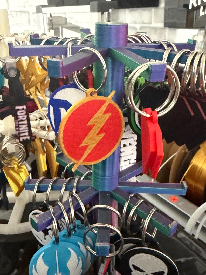 K&ouml;nnte beinhalten: Ein mehrfarbiger Ausstellungsst&auml;nder mit verschiedenen Schl&uuml;sselanh&auml;ngern. Die Schl&uuml;sselanh&auml;nger zeigen Logos und Designs, darunter ein rotes und goldenes Flash-Emblem. Der St&auml;nder hat eine zentrale S&auml;ule und horizontale Arme mit Metallringen.