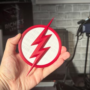 May include: A red and white circular emblem featuring a red lightning bolt. The emblem has a white center and a red border. The lightning bolt is a prominent design element, with sharp angles and a pointed tip. The emblem is held in a hand.