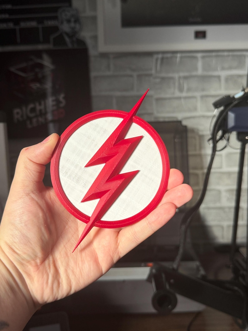 May include: A red and white circular emblem featuring a red lightning bolt. The emblem has a white center and a red border. The lightning bolt is a prominent design element, with sharp angles and a pointed tip. The emblem is held in a hand.