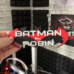 May include: A 3D printed sign featuring the black Batman bat-wing logo with a red accent layer. The text BATMAN & ROBIN is embossed in white lettering across the center of the emblem, creating a layered, multi-colored decorative display piece.
