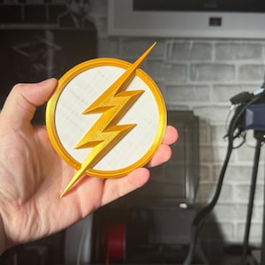 May include: A gold-colored lightning bolt emblem, set against a white circle, is framed by a gold border. The emblem is held in a hand, showcasing the 3D-printed design. The background is blurred, suggesting a workshop setting.